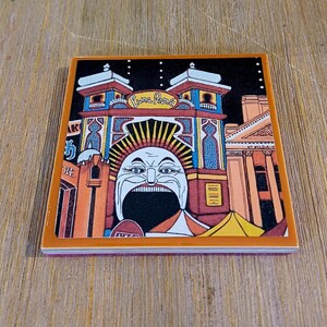 May include: A square ceramic coaster featuring a colorful illustration of the Luna Park entrance with its iconic wide-mouthed face, orange and blue architectural details, and the text Luna Park and St Kilda visible on the building facade.