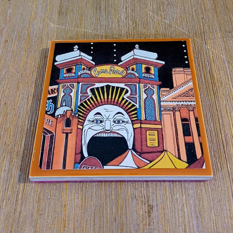 May include: A square ceramic coaster featuring a colorful illustration of the Luna Park entrance with its iconic wide-mouthed face, orange and blue architectural details, and the text Luna Park and St Kilda visible on the building facade.