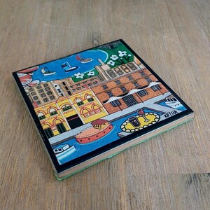Op de afbeelding: Een vierkante onderzetter met een kleurrijke illustratie van een waterkant met bakstenen gebouwen, een geel marktgebouw, boten op blauw water en een tafel met een glas wijn. De kunstenaarshandtekening Krysia is zichtbaar in de hoek.
