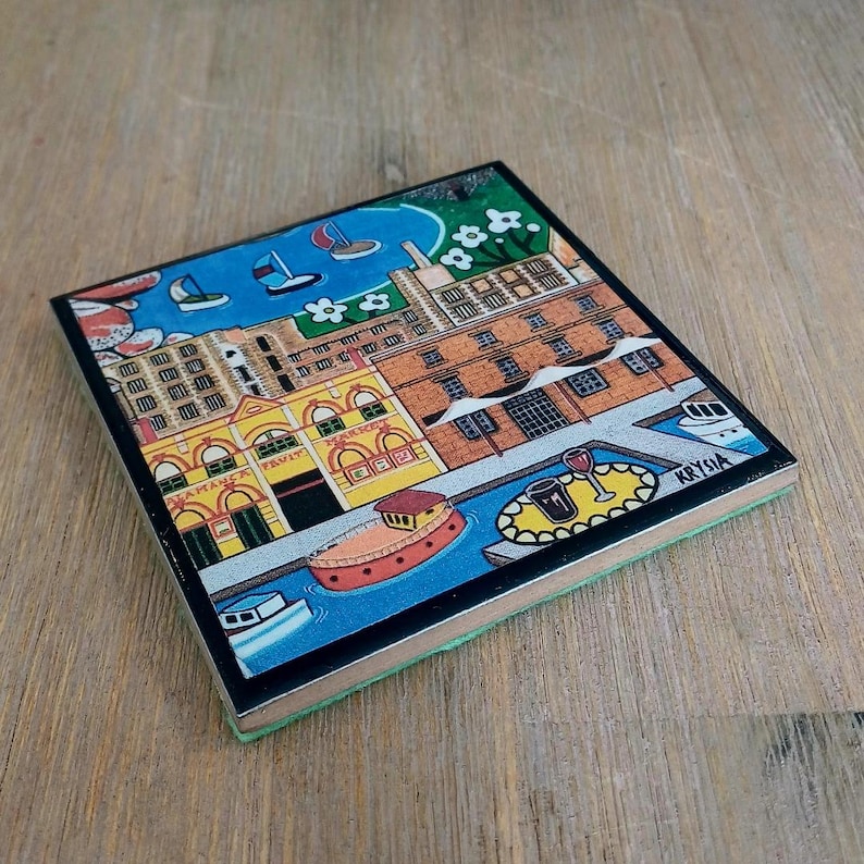 Op de afbeelding: Een vierkante onderzetter met een kleurrijke illustratie van een waterkant met bakstenen gebouwen, een geel marktgebouw, boten op blauw water en een tafel met een glas wijn. De kunstenaarshandtekening Krysia is zichtbaar in de hoek.