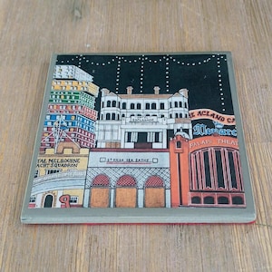 May include: A square ceramic coaster featuring a colorful illustration of St Kilda landmarks at night. The design includes the Hotel Esplanade, St Kilda Sea Baths, and the Palais Theatre with text labels against a dark sky with white dots.