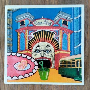 Puede incluir: Un azulejo cuadrado con una colorida ilustraci&oacute;n de la entrada del Luna Park con un rostro grande, un cubo verde con una pala, un plato con dos rebanadas de pan tostado y un tranv&iacute;a verde. La obra utiliza tonos vibrantes en azul, rojo y amarillo.