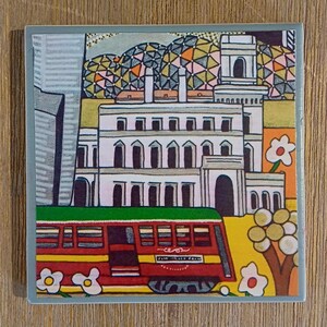 May include: A square ceramic tile featuring a colorful illustration of a red tram passing a white multi-story building. The background includes a geometric patterned dome, stylized trees with round leaves, and white flowers. The tram has text on its side.
