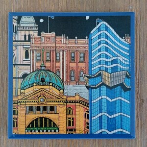 Op de afbeelding: Een vierkante keramische onderzetter met een kleurrijke illustratie van Flinders Street Station in Melbourne, Australi&euml;. De afbeelding toont het gele gebouw met de iconische groene koepel en klok, naast een moderne blauwe wolkenkrabber bij nacht.