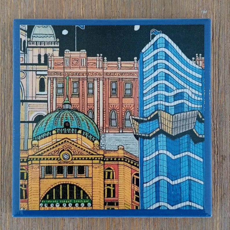Op de afbeelding: Een vierkante keramische onderzetter met een kleurrijke illustratie van Flinders Street Station in Melbourne, Australi&euml;. De afbeelding toont het gele gebouw met de iconische groene koepel en klok, naast een moderne blauwe wolkenkrabber bij nacht.