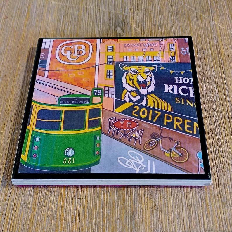 May include: A square coaster featuring a colorful illustration of a green North Richmond tram numbered 881, a tiger graphic, a bicycle, and a cafe table. The artwork includes text such as GB, Bryant & May, and 2017 PREM, set against a city street scene.