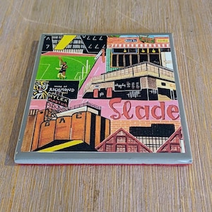 May include: A square coaster featuring a colorful graphic illustration of a cityscape with the text Home of Richmond, Nylex Plastics, GAE, and Slade. The artwork includes a green sports field, brick buildings, and architectural structures.