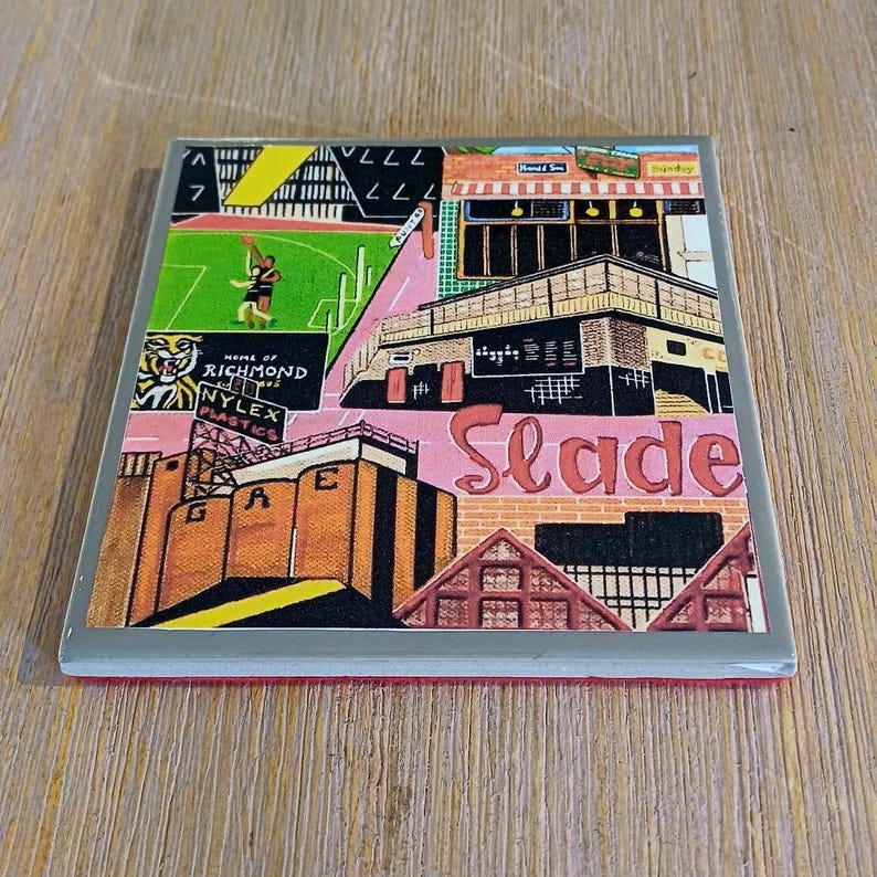 May include: A square coaster featuring a colorful graphic illustration of a cityscape with the text Home of Richmond, Nylex Plastics, GAE, and Slade. The artwork includes a green sports field, brick buildings, and architectural structures.
