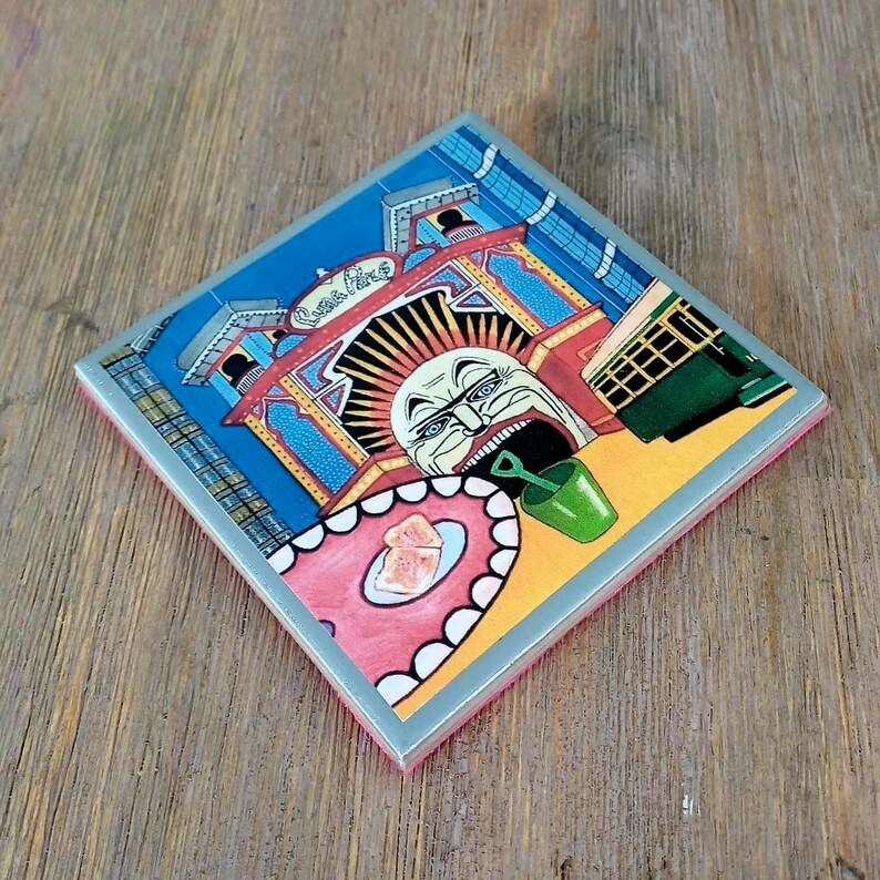 Puede incluir: Un posavasos cuadrado de cer&aacute;mica con una colorida ilustraci&oacute;n de la entrada del Luna Park que muestra un rostro estilizado, un cubo verde y un primer plano con forma de lengua rosa. La obra incluye el texto Luna Park en una fuente cursiva decorativa.