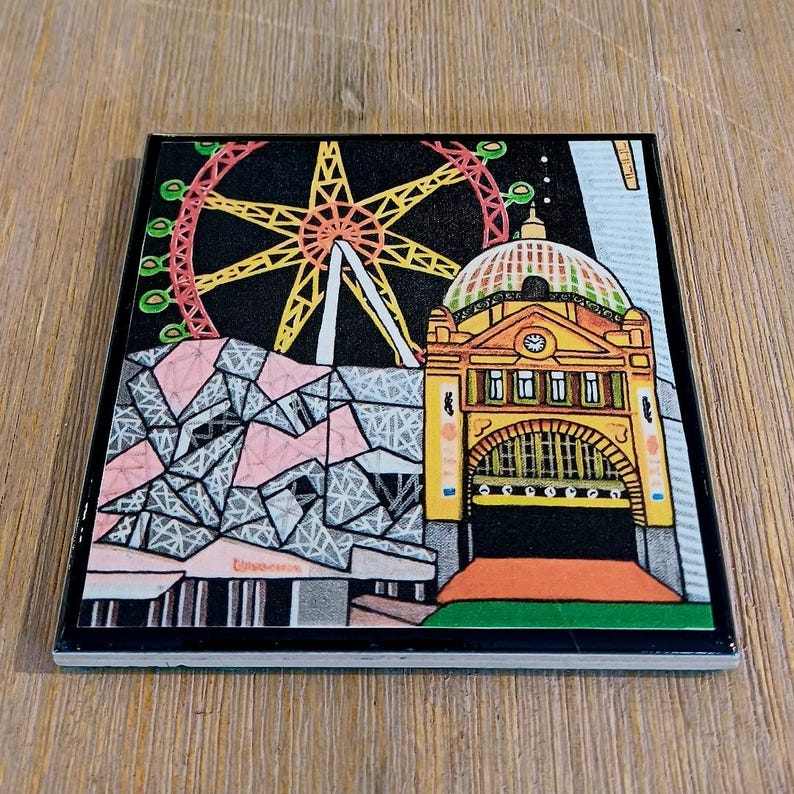 Puede incluir: Un posavasos cuadrado de cer&aacute;mica con una colorida ilustraci&oacute;n de monumentos de Melbourne, que incluye el edificio amarillo de la estaci&oacute;n de Flinders Street, una gran noria roja y una estructura de patr&oacute;n geom&eacute;trico sobre un cielo nocturno negro.