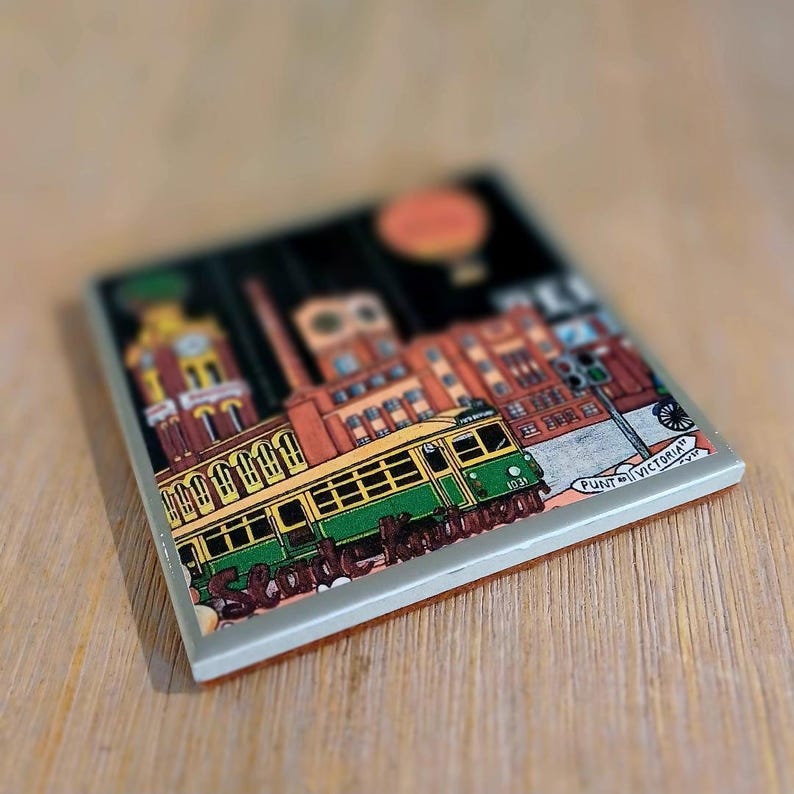May include: A square ceramic coaster featuring a colourful illustration of a green and yellow tram travelling past brick buildings. The text "PUNT RD" and "VICTORIA ST" is visible on a street sign, with "St Kilda" written in script below the tram.