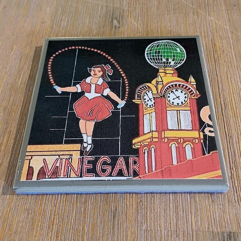 May include: A square coaster featuring a graphic illustration of a person jumping rope in front of a clock tower with a green globe on top. The word VINEGAR is printed in large red letters at the bottom against a black background with white lines.