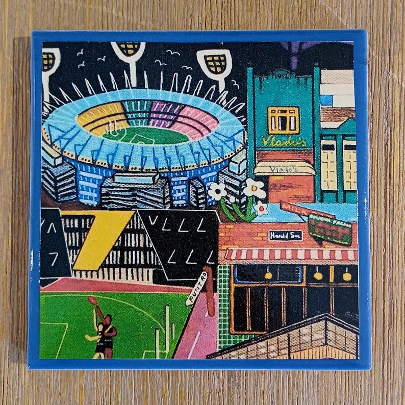May include: A square ceramic coaster with a blue border featuring a colorful illustration of a sports stadium, a green field with two figures playing, and city buildings. Text includes Vlado's, Herald Sun, and Punt Rd on the street signs.