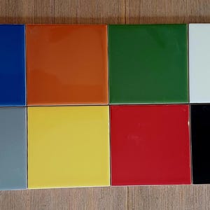 May include: A collection of eight square tiles arranged in two rows of four. The tiles feature a smooth, glossy finish in distinct solid colors including blue, orange, green, white, gray, yellow, red, and black, all displayed in a grid layout.