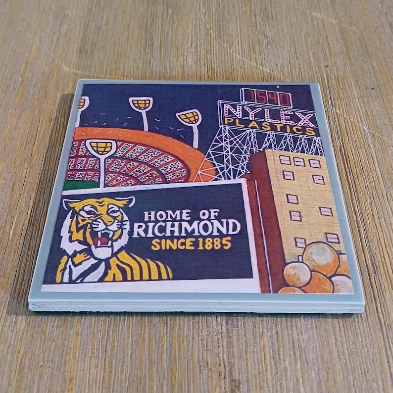 May include: A square coaster featuring a graphic illustration of a stadium, the Nylex Plastics sign, and a tiger mascot. The text reads Home of Richmond Since 1885 and Nylex Plastics. The design includes orange, yellow, white, and dark blue colors.