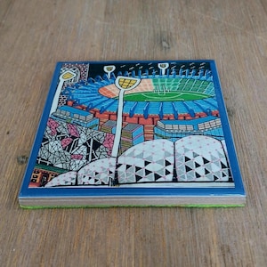 May include: A square ceramic coaster featuring a colorful graphic illustration of a sports stadium with a green field, blue seating, and tall light towers. The foreground displays a white architectural structure with black geometric patterns.