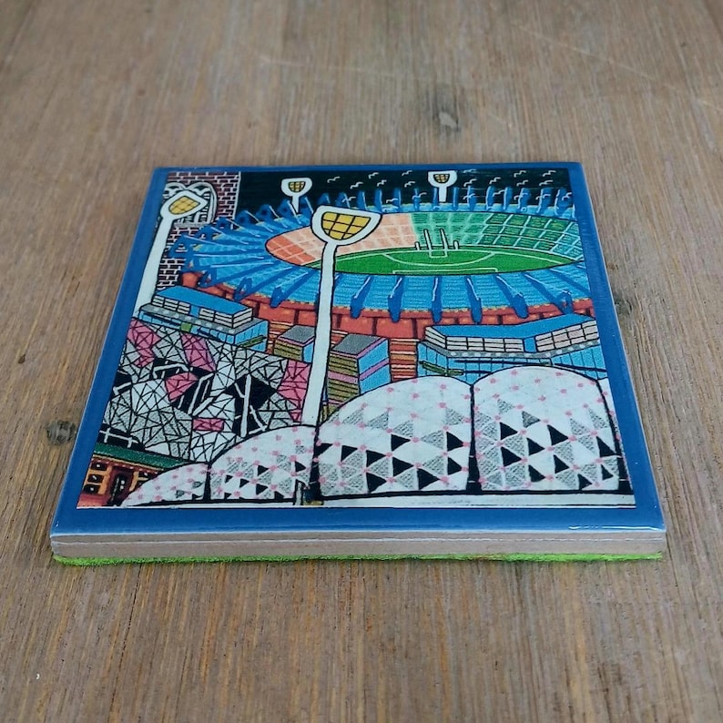 May include: A square ceramic coaster featuring a colorful graphic illustration of a sports stadium with a green field, blue seating, and tall light towers. The foreground displays a white architectural structure with black geometric patterns.