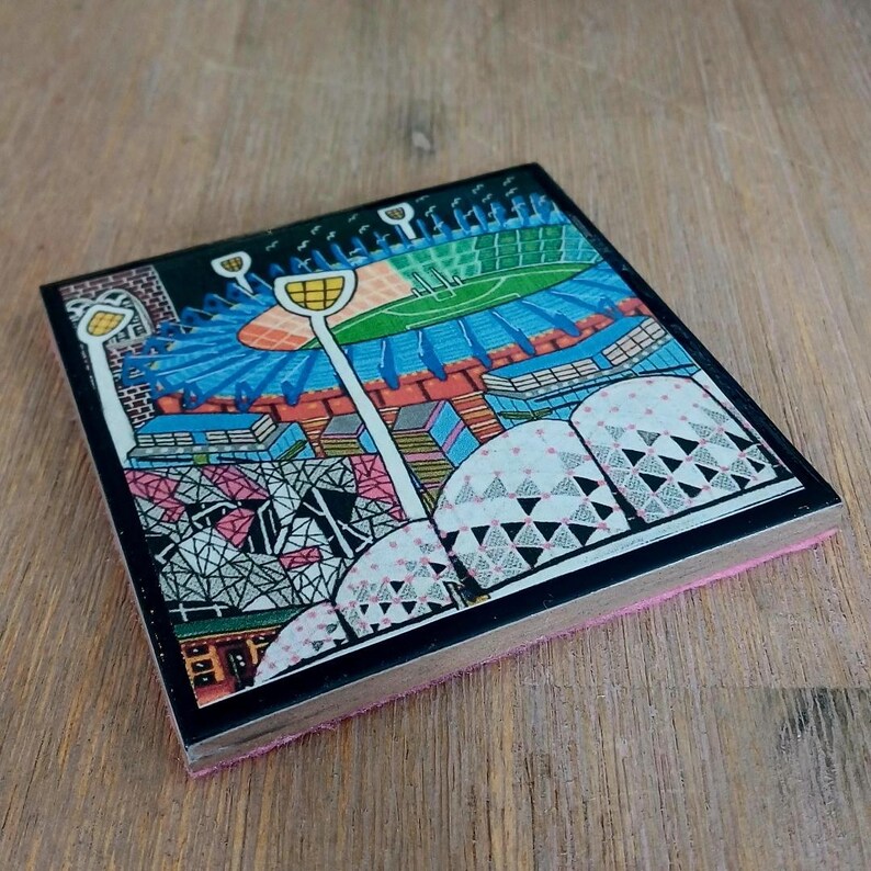 May include: A square coaster featuring a colorful graphic illustration of a stadium with a green field, blue seating, and tall white light poles. The design includes geometric patterns in pink, black, and white, with a black border and a pink base.