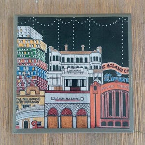 May include: A square coaster featuring a colorful illustration of the St Kilda skyline at night. The design includes the Hotel Esplanade, Palais Theatre, and St Kilda Sea Baths with text labels against a black sky with white dotted string lights.