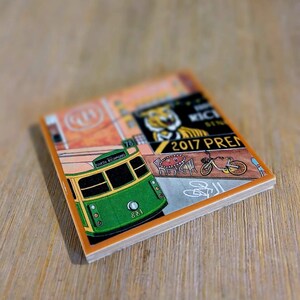 May include: A square ceramic coaster featuring a colorful illustration of a green and yellow tram labeled North Richmond 881, a bicycle, a cafe table with chairs, and text including 2017 PREM, all set against a warm orange and grey background.