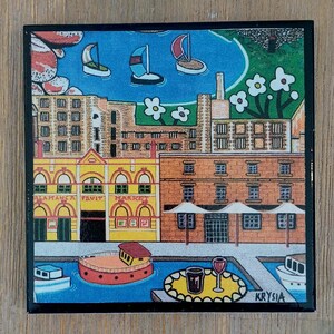 Op de afbeelding: Een vierkante decoratieve tegel met een kleurrijke illustratie van een stad aan het water. De afbeelding toont het Salamanca Fruit Market-gebouw, zeilboten op blauw water, bakstenen gebouwen, witte bloemen en een tafel met een wijnglas.