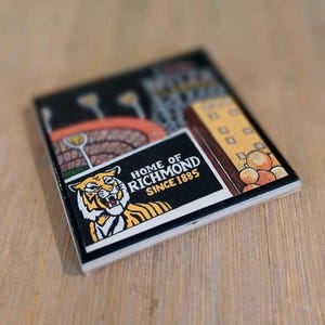 May include: A square ceramic coaster featuring a graphic illustration of a tiger head and the text HOME OF RICHMOND SINCE 1885. The design includes a stadium scene with orange seating and a yellow building structure on a black background.
