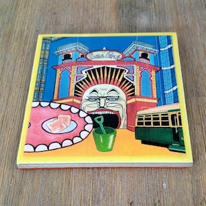 Puede incluir: Un posavasos cuadrado de cer&aacute;mica con una colorida ilustraci&oacute;n de la entrada del Luna Park que muestra una cara de payaso grande, un cubo verde con una pala, un plato rojo con dos tostadas y un tranv&iacute;a verde, todo enmarcado por un borde amarillo.