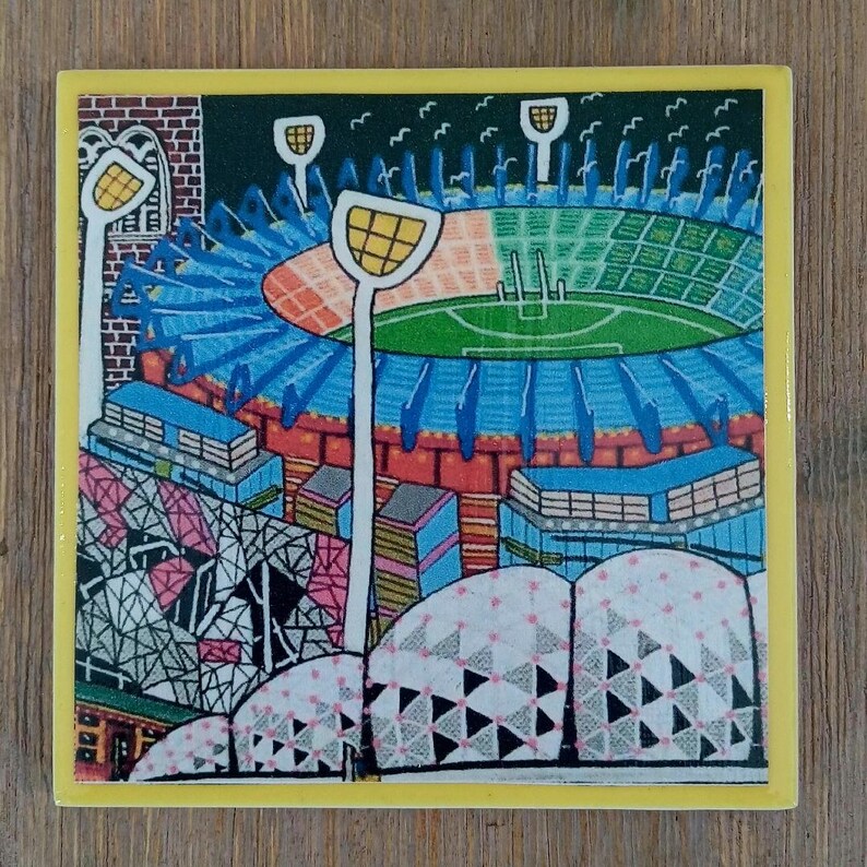 May include: A square ceramic tile featuring a colorful illustration of a sports stadium with a green field, blue seating sections, and tall white light towers. The artwork includes geometric patterns in the foreground and a yellow border.