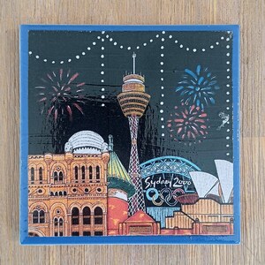 May include: A square decorative tile featuring a nighttime illustration of the Sydney skyline with the Sydney Tower, the Opera House, and the Sydney 2000 Olympic logo. The scene includes colorful fireworks against a black sky with white string lights.