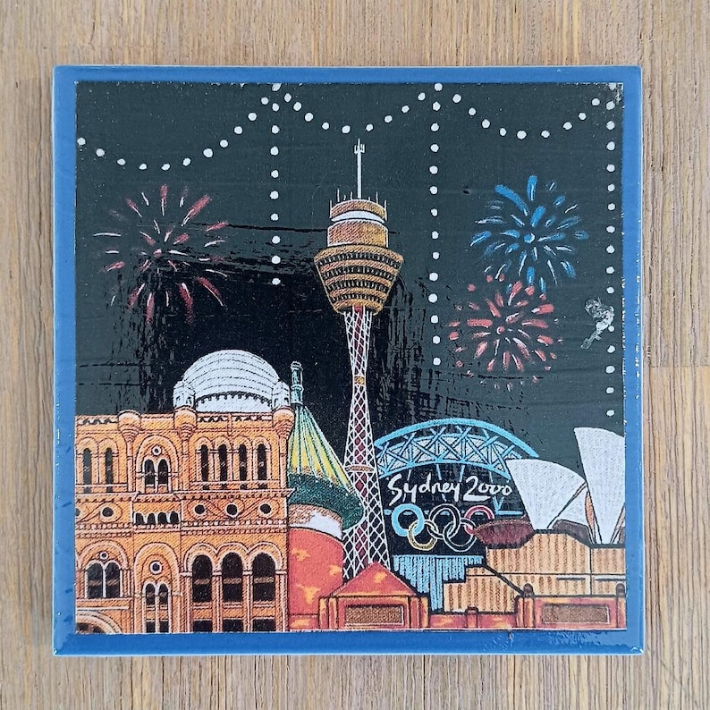 May include: A square decorative tile featuring a nighttime illustration of the Sydney skyline with the Sydney Tower, the Opera House, and the Sydney 2000 Olympic logo. The scene includes colorful fireworks against a black sky with white string lights.