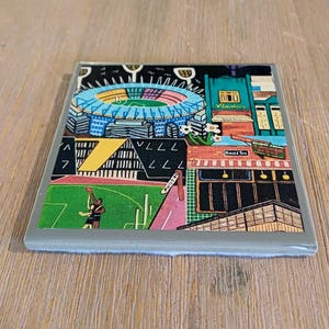 May include: A square ceramic coaster featuring a colorful illustration of a stadium with a blue roof, a green sports field with two figures, and surrounding city buildings with signs reading Vlade's, Herald Sun, and Punt Rd in a graphic style.