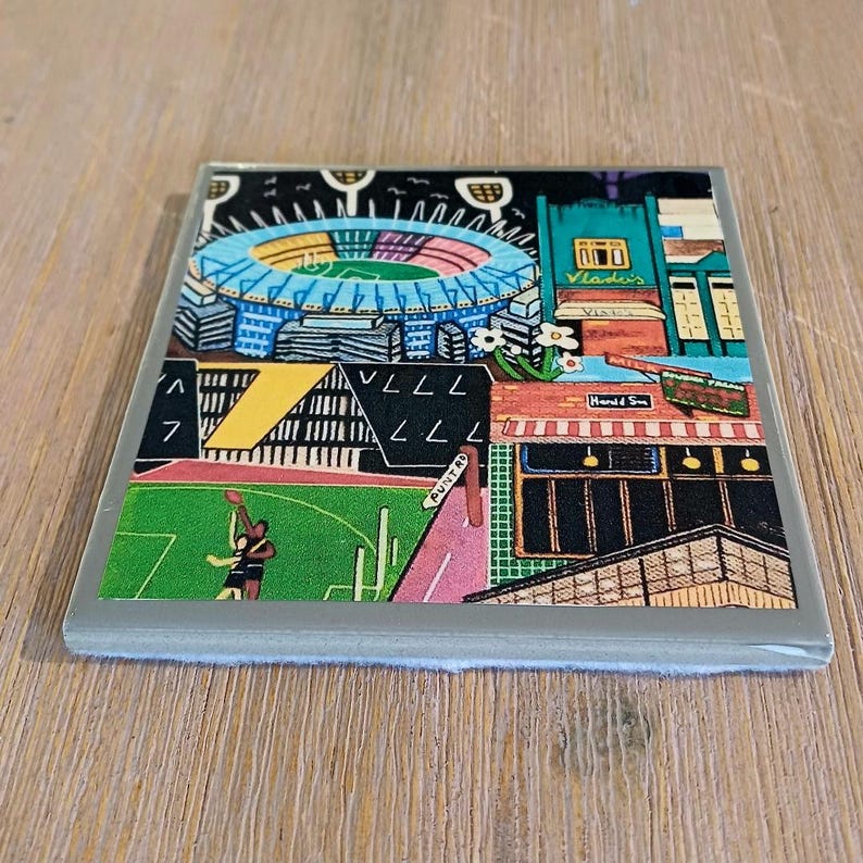 May include: A square ceramic coaster featuring a colorful illustration of a stadium with a blue roof, a green sports field with two figures, and surrounding city buildings with signs reading Vlade's, Herald Sun, and Punt Rd in a graphic style.