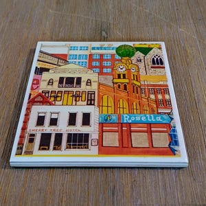 Peut inclure: Un dessous de verre carr&eacute; en c&eacute;ramique pr&eacute;sentant une illustration color&eacute;e d'une sc&egrave;ne de rue urbaine. L'&oelig;uvre repr&eacute;sente le Cherry Tree Hotel et un b&acirc;timent avec une tour d'horloge &eacute;tiquet&eacute; Rosella, dans des tons de rouge, jaune, bleu et blanc.