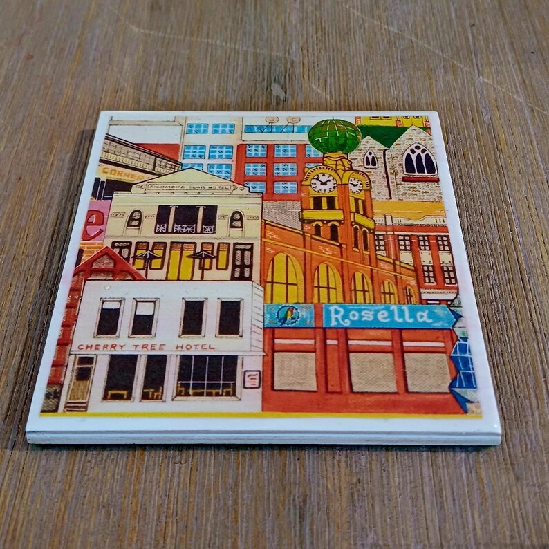 Peut inclure: Un dessous de verre carr&eacute; en c&eacute;ramique pr&eacute;sentant une illustration color&eacute;e d'une sc&egrave;ne de rue urbaine. L'&oelig;uvre repr&eacute;sente le Cherry Tree Hotel et un b&acirc;timent avec une tour d'horloge &eacute;tiquet&eacute; Rosella, dans des tons de rouge, jaune, bleu et blanc.