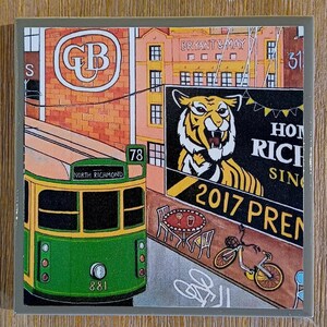 May include: A square ceramic tile featuring a colorful illustration of a green and yellow tram labeled North Richmond 78, a brick building with a Bryant & May sign, a tiger graphic on a billboard, a small outdoor cafe table, and a bicycle.