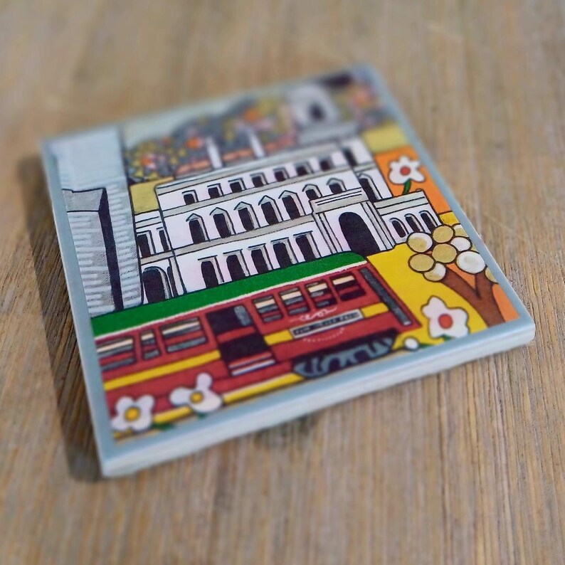 May include: A square ceramic tile featuring a colorful illustration of a white multi-story building with arched windows and a red streetcar passing in front. The scene includes stylized flowers, a yellow background, and a gray structure on the left side.
