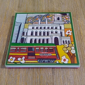 May include: A square ceramic coaster featuring a colorful illustration of a white building with arched windows, a red streetcar in the foreground, and stylized flowers. The design includes a geometric patterned sky and a vibrant yellow ground.