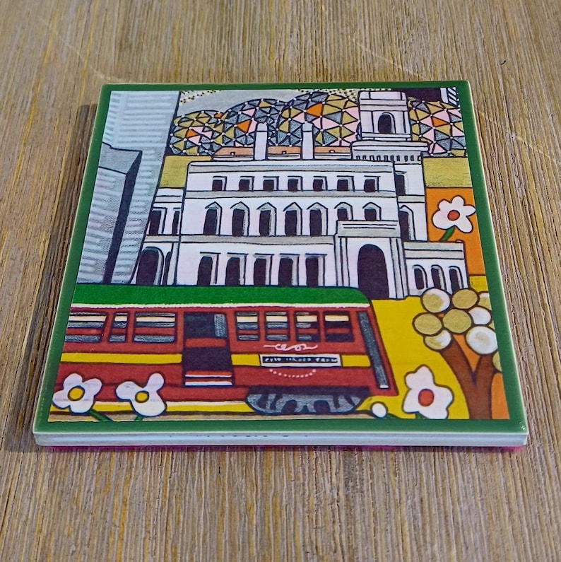 May include: A square ceramic coaster featuring a colorful illustration of a white building with arched windows, a red streetcar in the foreground, and stylized flowers. The design includes a geometric patterned sky and a vibrant yellow ground.