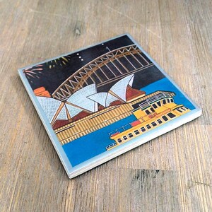 May include: A square coaster featuring a colorful illustration of the Sydney Harbour Bridge and Opera House at night. A yellow ferry boat travels across the blue water in the foreground, while fireworks are visible in the dark sky above the bridge.