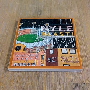 May include: A square ceramic coaster featuring a colourful graphic illustration of a stadium and urban buildings. The design includes the text NYLE PLASTI, BRYANT & MAY, and the number 3121 displayed twice in a stylised font against a dark background.