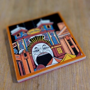 May include: A square ceramic coaster featuring a colorful graphic illustration of a carnival entrance with a large, stylized face with a wide-open mouth at the center. The design includes blue, orange, and black tones with architectural details.