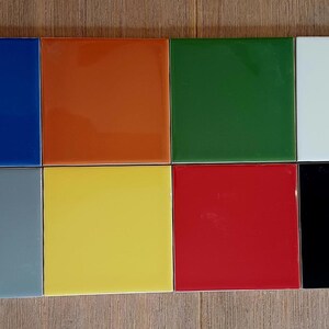 May include: A collection of eight square tiles arranged in two rows of four. The tiles feature a smooth, glossy finish in distinct solid colors including blue, orange, green, white, gray, yellow, red, and black, all displayed in a grid layout.