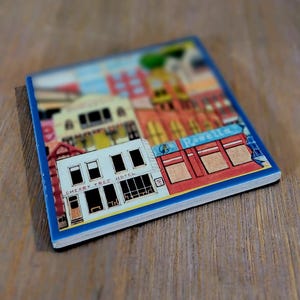 Peut inclure: Un dessous de verre carr&eacute; pr&eacute;sentant une illustration color&eacute;e d'une sc&egrave;ne de rue avec des b&acirc;timents. Le b&acirc;timent blanc au premier plan porte l'inscription Cherry Tree Hotel, et un b&acirc;timent rouge voisin affiche le texte Rosella sur un panneau bleu.