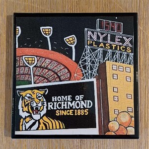 May include: A square coaster featuring a graphic illustration of a stadium with floodlights, a Nylex Plastics sign with the number 1540, a yellow building, and a tiger head graphic with the text Home of Richmond Since 1885 on a black background.