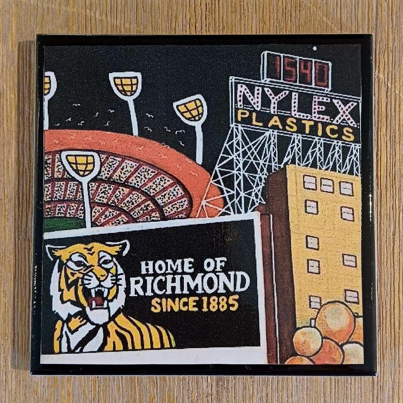 May include: A square coaster featuring a graphic illustration of a stadium with floodlights, a Nylex Plastics sign with the number 1540, a yellow building, and a tiger head graphic with the text Home of Richmond Since 1885 on a black background.