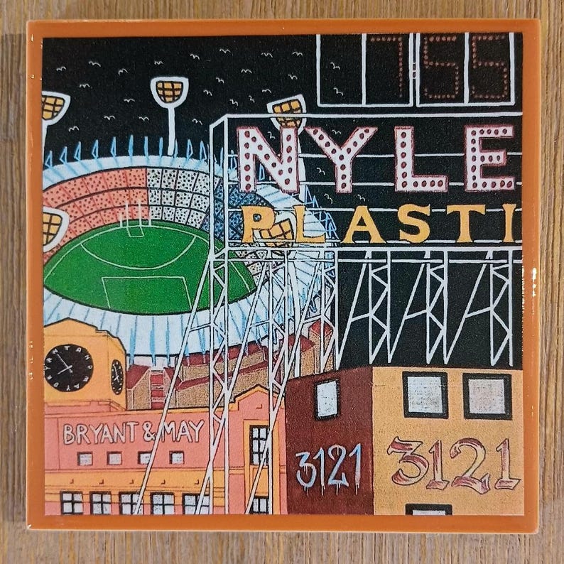 May include: A square ceramic tile featuring a colourful illustration of a stadium with a green field, floodlights, and a scoreboard. The text NYLE PLASTI, BRYANT & MAY, and 3121 are visible. The tile has a thick orange border and a glossy finish.
