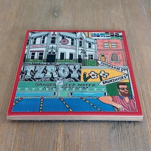 May include: A square ceramic coaster with a red border featuring a colorful illustration of a street scene with buildings, the text FITZROY, Johnson St, Brunswick St, and Danger Deep Water, and a man with a beard and sunglasses in a striped shirt.