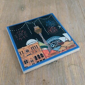 May include: A square ceramic coaster featuring a colorful illustration of the Sydney 2000 Olympic skyline at night. The design includes the Sydney Tower, the Opera House, and fireworks against a black sky with the text Sydney 2000 visible.