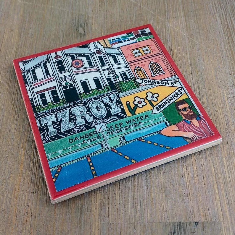 May include: A square ceramic coaster with a red border featuring a colorful illustration of a city street scene. The artwork includes buildings, street signs labeled Johnson St and Brunswick St, the text Fitzroy, and a person with a beard.