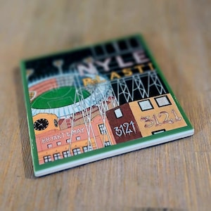May include: A square ceramic coaster featuring a colourful illustration of a stadium and industrial buildings. The text reads NYLE, PLASTI, BRYANT & MAY, and 3121. The design includes a clock tower, floodlight towers, and a green field under a dark sky.